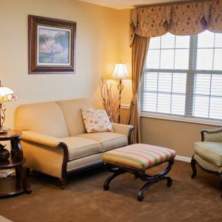 A cozy living room with a beige sofa, armchair, and a small footstool, featuring warm lighting and large windows with curtains.