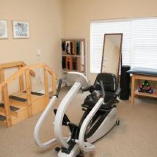 A rehabilitation therapy room featuring an elliptical machine, wooden steps, a mirror, and shelves beside a window.