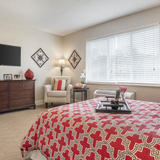 A cozy bedroom with a red-patterned bedspread, a chair near the window, and a television mounted on the wall.