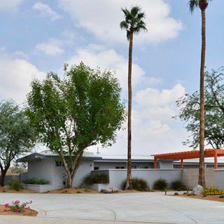 A mid-century modern house with palm trees and a cloudy sky backdrop.