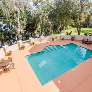 A tranquil outdoor swimming pool area with patio seating surrounded by lush greenery on a sunny day.
