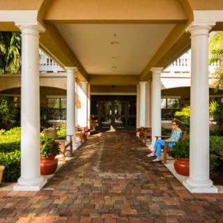A covered passageway with columns and a brick walkway, featuring outdoor seating and surrounding garden plants.