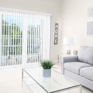 A modern living room with a comfortable sofa and glass coffee table, featuring natural light from a large sliding glass door with vertical blinds.