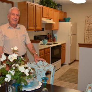 A man standing in a well-decorated kitchen with wooden cabinets and a dining area featuring a floral centerpiece.