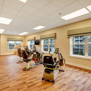 A bright and spacious fitness room with exercise equipment, including treadmills and stationary bikes, set against large windows allowing natural light.