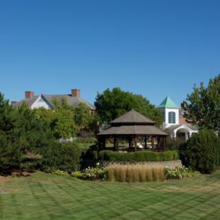 A scenic view of a garden with a gazebo, surrounded by trees and a well-maintained lawn, with a house in the background under a clear blue sky.