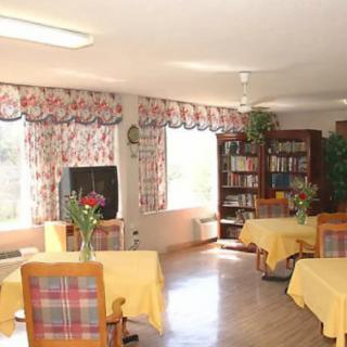 A cozy dining area with tables and chairs, adorned with yellow tablecloths, floral curtains, and a bookshelf.