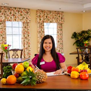 A woman smiling while writing at a table filled with various fruits in a bright kitchen.