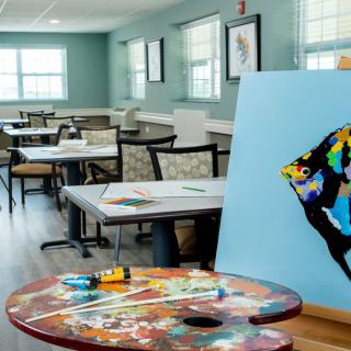 A brightly lit art room featuring a colorful fish painting on an easel and a paint palette on the table.
