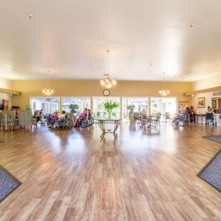 A spacious and bright community room in a senior living facility, featuring wooden flooring and elegant decor.