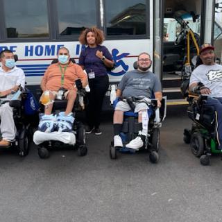 A group of individuals in wheelchairs, accompanied by a caregiver, gathered outside a transportation vehicle designed for accessibility.