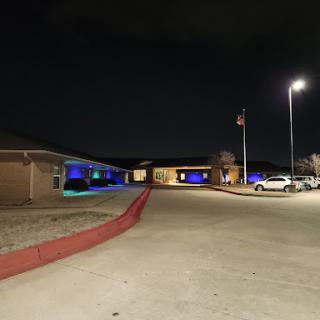A nighttime view of a building illuminated with blue lights, featuring a parking lot and an American flag.