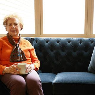 An elderly woman sits comfortably on a tufted sofa, enjoying the warm natural light coming through the windows.