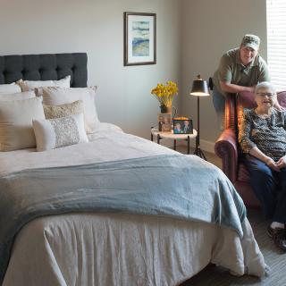 A cozy bedroom with a neatly made bed and two people sitting on a chair beside it.
