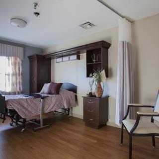 A well-furnished hospital patient room featuring a bed, chair, and natural light.