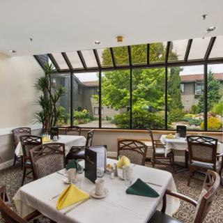 A bright dining area with large windows overlooking a garden, featuring neatly set tables and chairs.