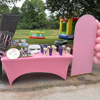 A beautifully arranged pink table setup for an outdoor event, complete with decorations and fun activities in the background.