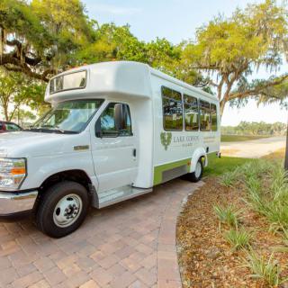 A white shuttle bus parked on a paved path, surrounded by trees and greenery.