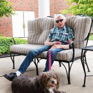 A man wearing sunglasses sits casually on an outdoor bench with a dog by his side.