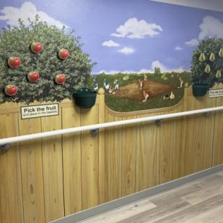 A mural featuring apple trees and a scenic landscape with handrails along the wall, possibly in a nursing home.