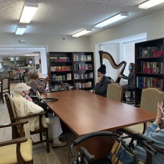 A peaceful library setting with seniors gathered around a table, surrounded by shelves of books and a harp in the corner.