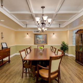 A well-designed dining room featuring a large wooden table with chairs, illuminated by an elegant chandelier. The room is adorned with wall art and decorative plants, accentuating the classic ceiling design.