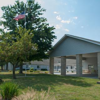 A serene view of a building with an American flag waving in the breeze, surrounded by trees and green grass under a clear blue sky.