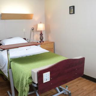 A clean and simple hospital room featuring an adjustable bed with a green blanket, a bedside lamp, and minimalistic decor.