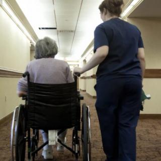 A nurse assists an elderly person in a wheelchair down a hallway.