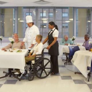 A chef and a server assisting elderly residents in a nursing home dining hall.