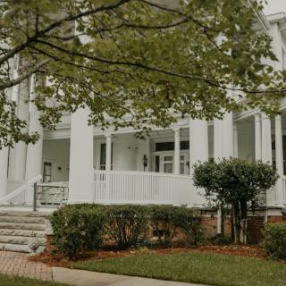 A white house with a porch, surrounded by trees and a neatly trimmed garden.