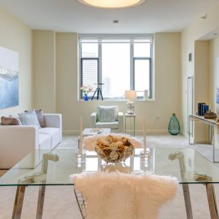 A modern and minimalistic living room with natural light streaming through the window, featuring a glass table and neutral-toned furniture.