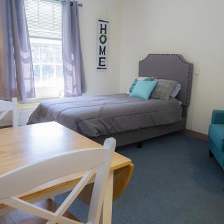 A cozy bedroom with a single gray-bedded setup, accompanied by a wooden table and chair next to a blue sofa, under soft natural light from the window.