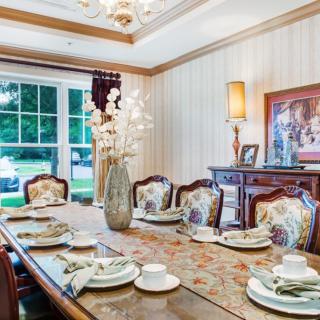 An elegantly furnished dining room featuring a well-set table with ornate chairs, a decorative vase centerpiece, and ambient lighting from a chandelier overhead.