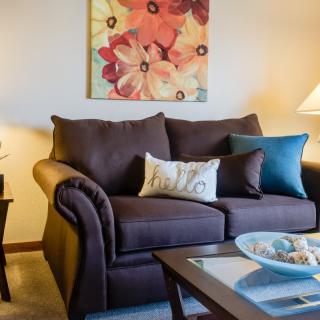 A cozy living room with a brown sofa, colorful cushions, and a floral painting on the wall.