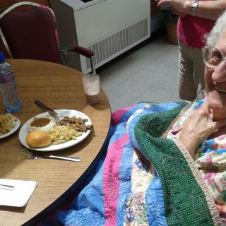 An elderly woman sitting at a dining table with a meal and wrapped in a blanket.