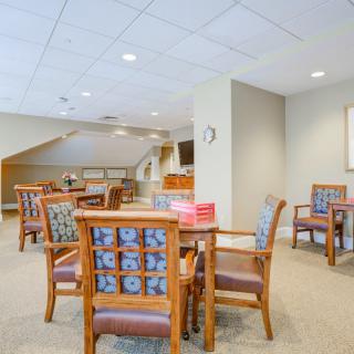 A comfortable seating area with wooden chairs and tables, featuring a carpeted floor and bright lighting.