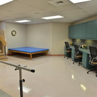 A rehabilitation therapy room featuring a blue exercise mat, support bar, and workstations.