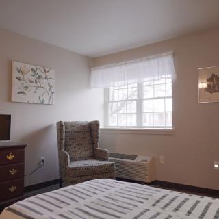 A cozy bedroom with a bed, armchair, and framed artwork near a bright window.