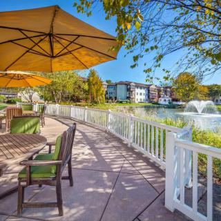 A scenic outdoor patio with seating and umbrellas, overlooking a lake with a fountain.