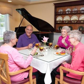 A group of elderly people enjoy a meal together at a dining table with a piano in the background.
