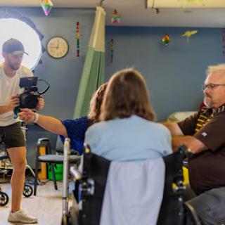 A camera operator films an interview in a care facility, capturing interactions among residents.