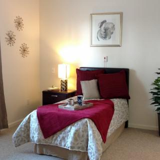 A cozy bedroom with a single bed, featuring red and beige decor, a lamp on the nightstand, and wall art.