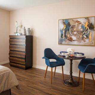 A modern bedroom featuring a cozy dining area with two blue chairs, a small round table, and abstract artwork on the wall.