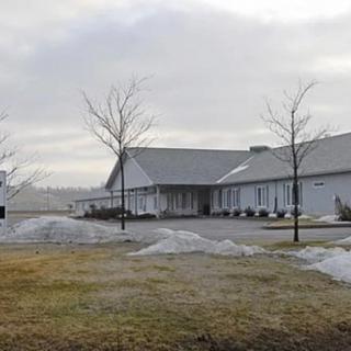 A large building with a grey roof stands on an overcast winter day, surrounded by patches of snow and barren trees.
