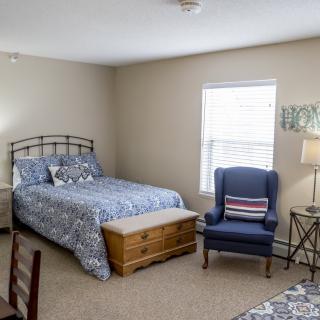 A cozy bedroom with a metal bed frame, patterned bedding, an armchair, and a window allowing natural light.