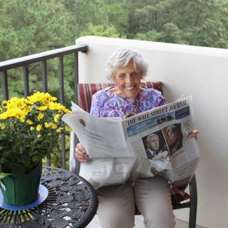 An elderly woman relaxes on a balcony while reading a newspaper, surrounded by flowers.
