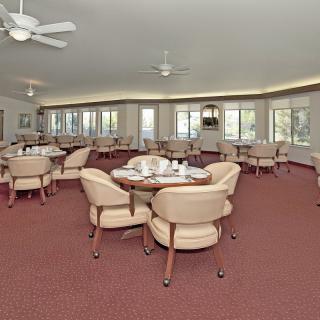 A spacious dining room with multiple tables and chairs, featuring large windows and ceiling fans.