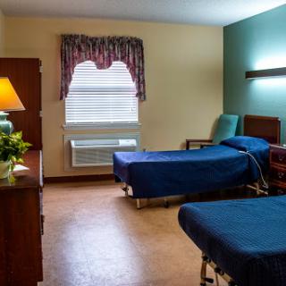 A view of a hospital room featuring two beds, a nightstand, and a window with curtains.