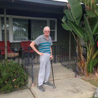 An elderly man stands on the porch of his home, using a cane for support.